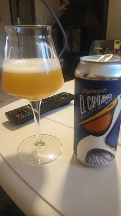 Ett glas med ljus öl och en ölburk med Stigbergets El Capitano på ett bord, fjärrkontroll i bakgrunden.