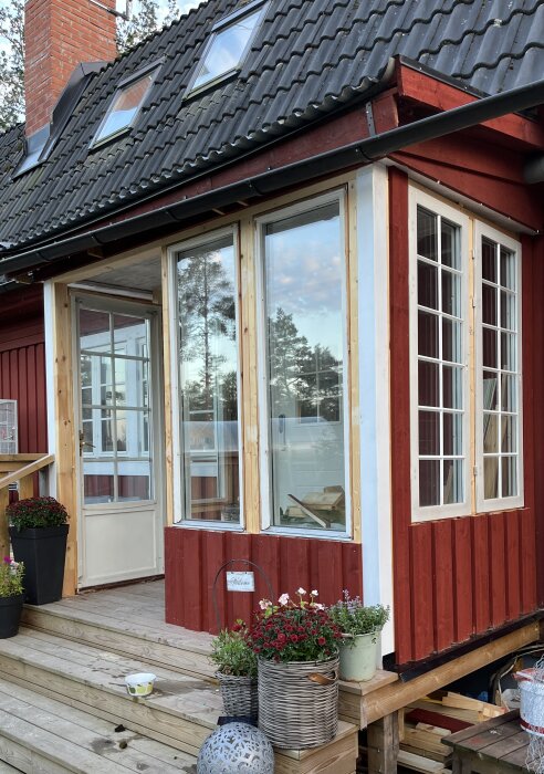 Röd träfasad, veranda med trappa, planterade blommor i krukor. Huset har fönster och hängränna, arbetsprojekt pågår.