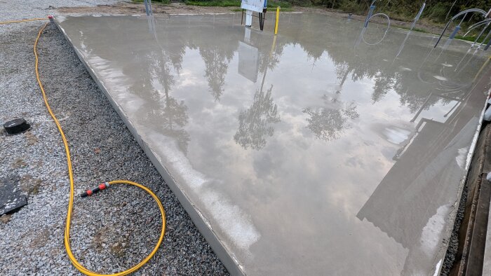 Nylagd betongplatta med vattenspegel, en gul trädgårdsslang på grus bredvid; reflektioner av träd och himmel syns på ytan.