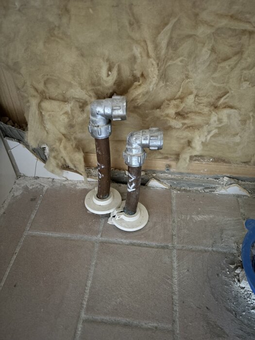 Två inkommande vattenrör som sticker upp ur betongplattan, med vinklade kopplingar och isolering i bakgrunden. Golvet är kaklat.