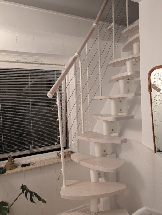 Vit spiraltrappa med trästeg och vajerräcke, placerad inomhus nära ett fönster.
