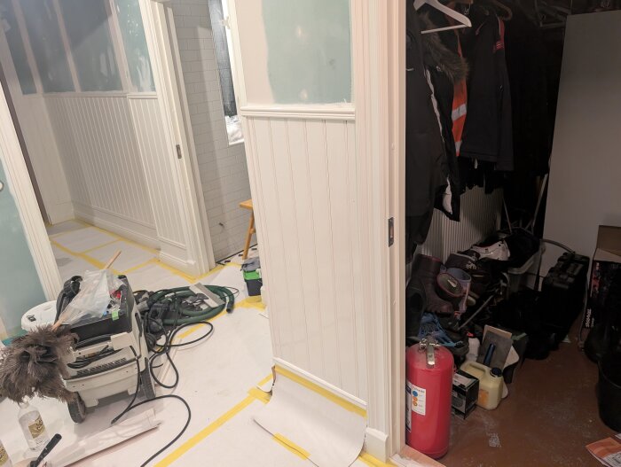 Renovering av rum med målad pärlspont, verktyg och material på golvet, golvet täckt med papper, och ett öppet förråd med kläder och skor.