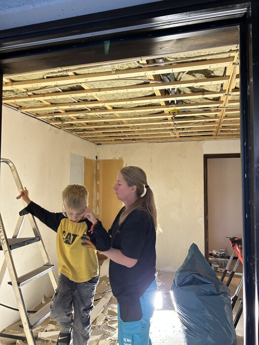 Barn och vuxen vid stege i rum under renovering, synligt exponerat tak och verktyg på golvet.