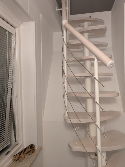 Spiraltrappa med ett räcke av trä och metallvajrar, visar fäste i väggen och girlande steg med synliga skruvar, belägen nära ett fönster med jalusi.