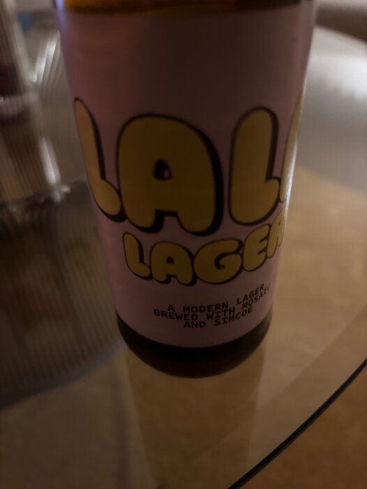 Flaska med etikett "Lal Lager", modern lager, bryggd med Mosaic och Simcoe, på ett bord.