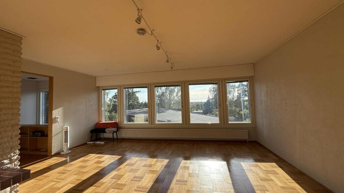 Tomt vardagsrum med parkettgolv, stora fönster med utsikt mot träd, och ett element samt en stol vid sidan.