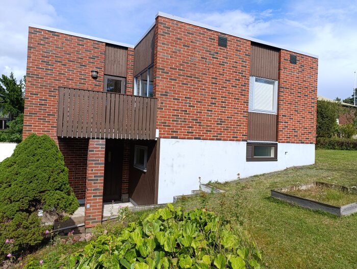 Tegelhus med trädetaljer, balkong och grön trädgård med buskar i förgrunden under en blå himmel.