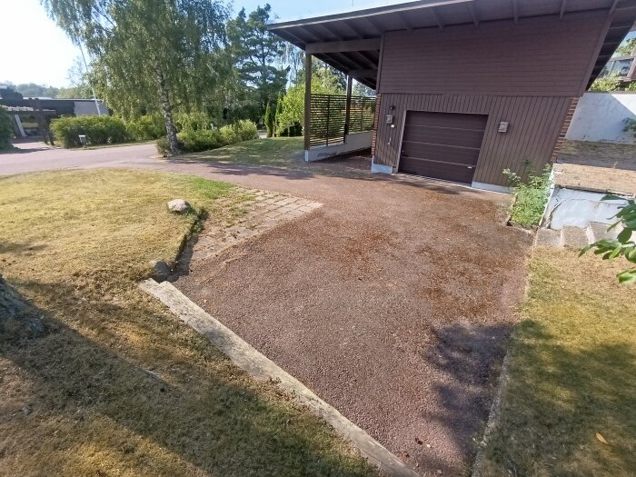 Uppfart med grus och gräs, garage intill, soligt väder, träd och buskar i närheten, potentiellt arbetshandlingar att utföra.