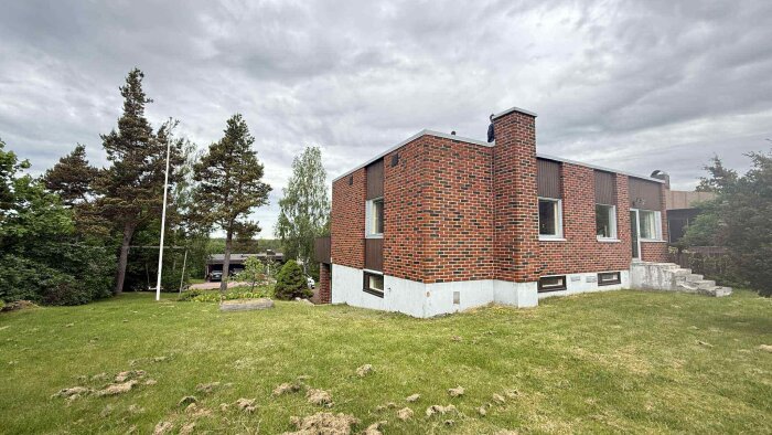 Tegelhus med platt tak, omgiven av en grön gräsmatta och träd, med en person synlig på taket under molnig himmel.