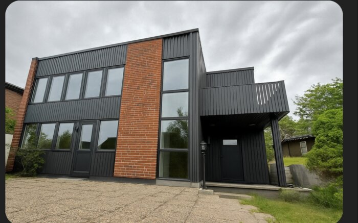 Modern byggnad med svart fasad och tegeldetaljer, stor balkong planeras med tonat glasräcke enligt text.