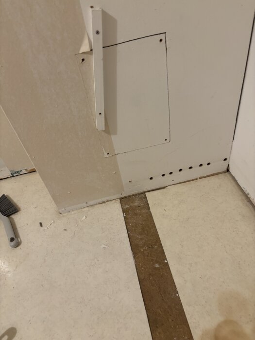 Väggpanel med trapplock, skruvar och en list syns. Golvet har trädetaljer, och en borste ligger på sidan.