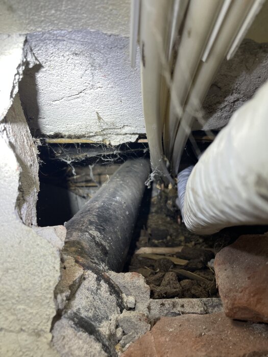 Hål i husvägg med exponerade rör och elkablar, omgivna av tegel och byggmaterial, i en källare från tidigt 1900-tal.