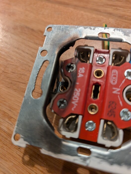 Bränt eluttag med metallram och elkablar synliga, märkning "ELKO" och värdena "16A" och "250V" på röd plast.