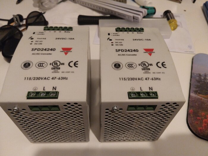 Två 24VDC-10A strömförsörjningsenheter, modell SPD24240, för verkstadsbelysning, ligger på ett bord med diverse små föremål runt omkring.