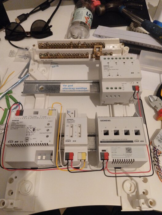KNX-komponenter uppsatta på arbetsbord, med bl.a. flera Siemens-moduler anslutna med kablar. Verktyg och flaskor i bakgrunden.