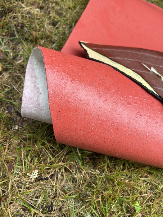 Röda golvmattor som har rivits ut ligger på en gräsmatta, med ett synligt vitt innermaterial i en av mattorna.