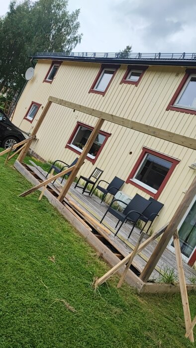 Ofärdig altan med golv och stolpar, saknar räcken och tak, framför ett gult hus. Flera stolar står på altanen, gräs i förgrunden.