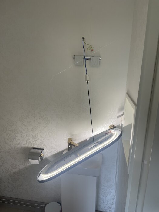 Nedfallet badrumsspegel över handfat med elektriska kablar synliga, delvis lossad väggmontering, toalettrullehållare och mönstrad tapet i bakgrunden.