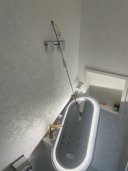 Spegel har lossnat från väggen ovanför ett handfat, elektriska kablar hänger löst, spegeln stöttas av ett snett stående föremål.