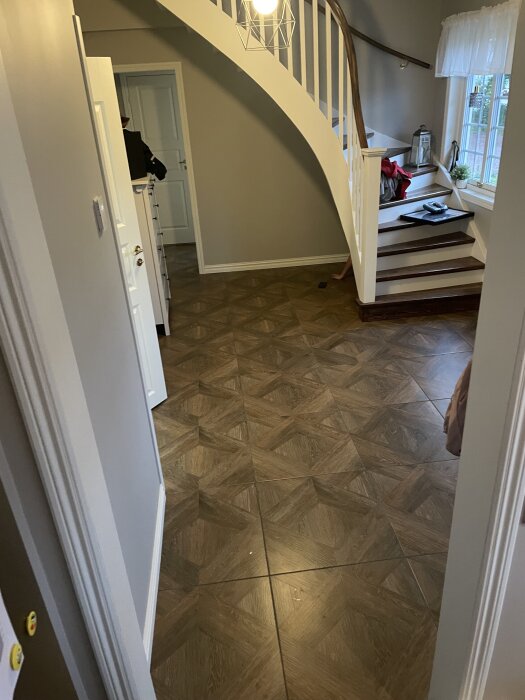 Trappa i en hall med parkettgolv och en designlampa i taket, sett från en dörröppning.