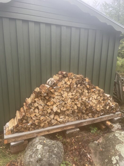 Uppstaplad ved mot en grön bod, placerad på ett upphöjt golv av trall och 45x70-reglar på betongblock, intill en stor sten.