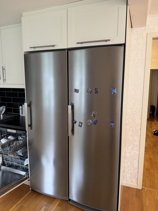 Rostfria kylskåp med magneter, omgivna av vita IKEA-överskåp och en diskmaskin med öppen lucka till vänster.