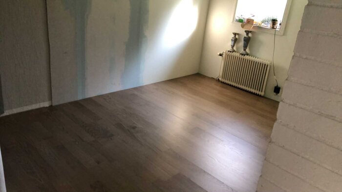 Renoverat rum med nylagt trägolv, en radiator under fönster och målad vägg i olika nyanser.