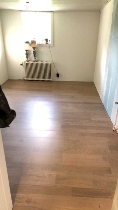 Ett rum med ett trägolv, ett element under ett fönster och en ofärdig vägg redo för renovering och eldragning.