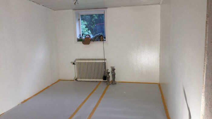 Tomt rum med vita väggar och grått golv, element under fönster och tejp vid kanterna.