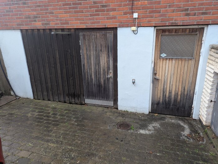 Trägarage med två dörrar, tegelvägg ovanpå. Blött område vid ena dörren, antyder vattenläckage. Brunn på marken framför.
