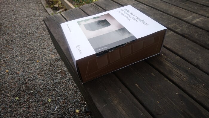 Trasig kartong med Zaptec Go 2-förpackning på träbord vid grusgång, texten "Asphalt Black" synlig.