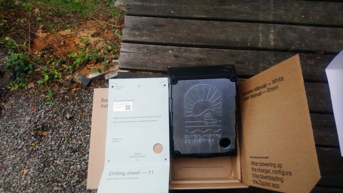 Kartongöppning med "zaptec go 2"-installationsdelar och en borrmall ligger på ett träbord utomhus, med text och logotyper synliga.