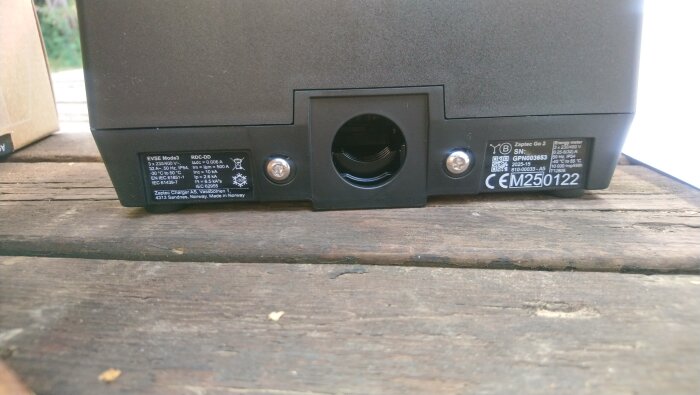 Baksidan av en laddboxmodell "Zaptec Go 2" med etiketter och skruvar synliga, placerad på ett trädäck.