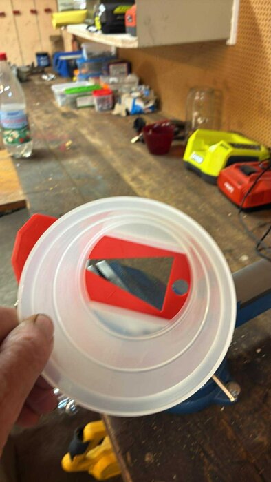 En hand håller en rund plastbehållare med ett rött handtag och en spegelliknande yta; okänd funktion.