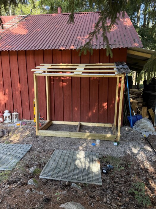 En enkel trästomme byggd av återvunnet virke, placerad framför en röd byggnad med plåttak, stående på grusmark.