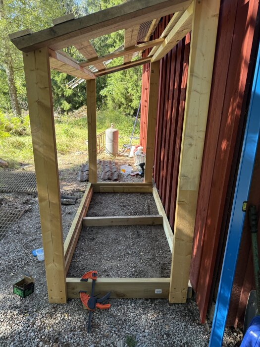 Träram för byggprojekt, konstruerad av återvunnet virke, står lutad mot en röd byggnad med skog i bakgrunden.