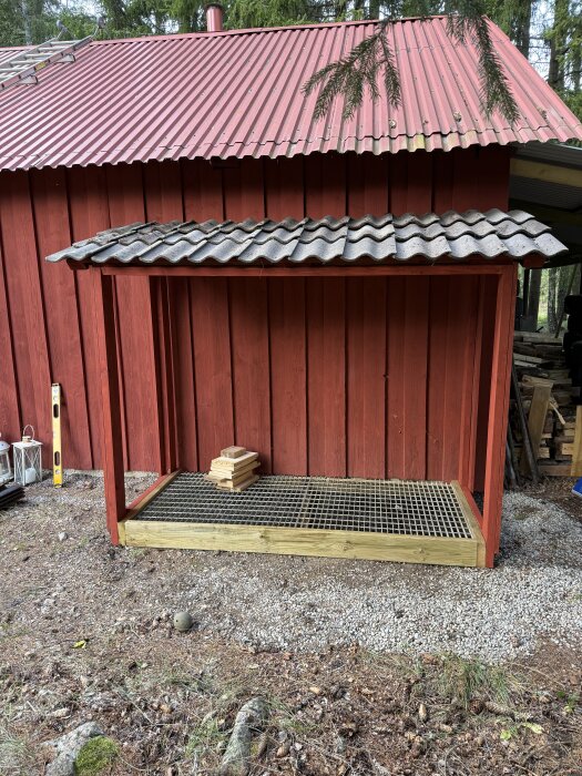 En enkel träkonstruktion med tak och gallerbotten, byggd mot en röd trävägg med vågformat plåttak.