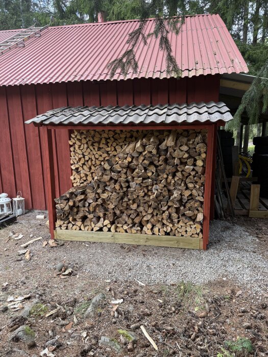 Vedstapel under enkel träkonstruktion bredvid röd byggnad.
