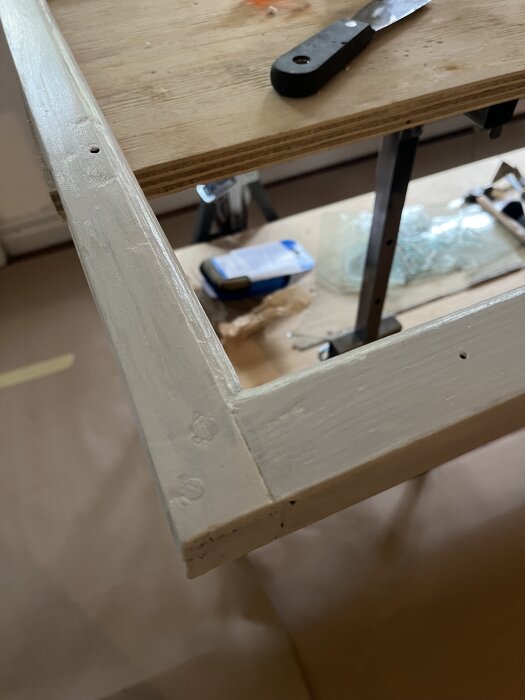 Fönsterbåge målad med antikvit linoljefärg, stående på arbetsbänk med verktyg och material i bakgrunden.