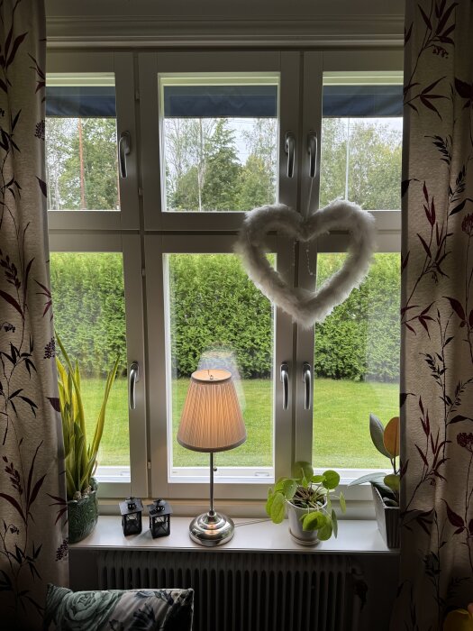 Fyra vita flaggfönster med ett hjärtformat hänge, en bordslampa och växter på fönsterbrädan, utsikt mot trädgård.