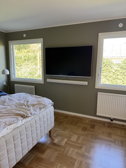 Sovrum med uppsatt TV och soundbar på väggen, säng till vänster och två fönster med utsikt över trädgården.