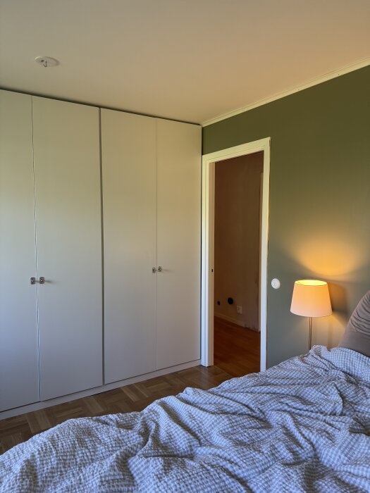 Sovrum med grå garderober, grön vägg, lampa och säng med ljus filt.