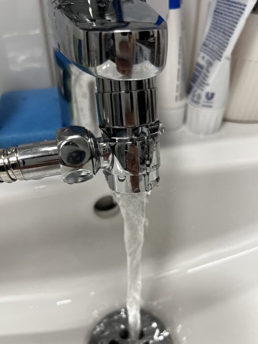 Vatten rinner från en kromad tvättblandare med gängade kopplingar, omgiven av verktyg och tandvårdsprodukter.