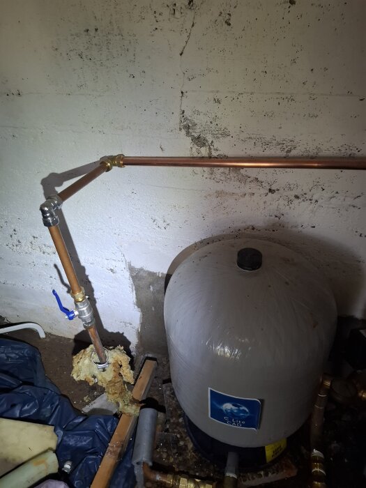 Vattenbehållare och kopparrör mot en vit vägg i en källare, med isolering runt ett rör.