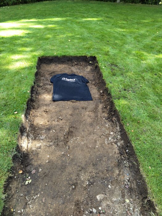 Grävd grop i gräsmatta med en svart tröja från byggahus.se placerad i botten.