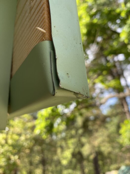 Närbild av takets kantfals med grönt plåtmaterial och trädetaljer, mot en bakgrund av suddiga träd.