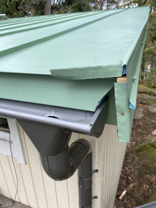 Falsat plåttak med grön kant vid husets hörn, intilliggande hängränna och stuprör i metall.