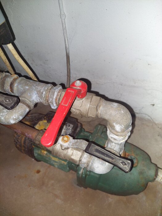 Vattenpump med rörkopplingar och röda samt svarta handtag på ventiler, placerad mot en vit vägg.