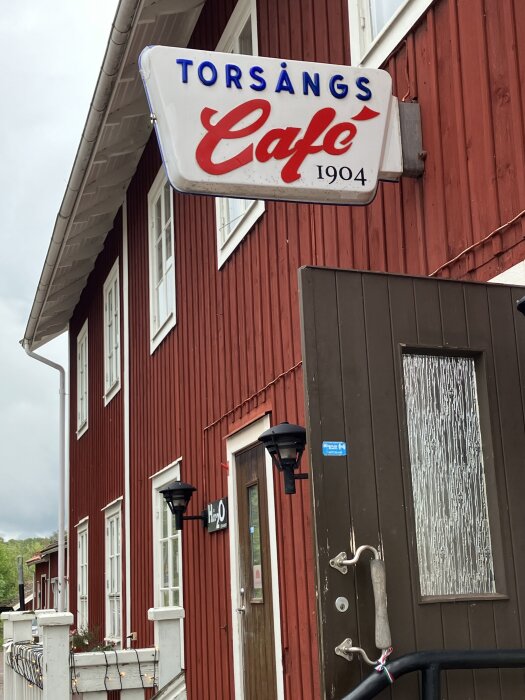 Rött trähus med skylt: "Torsångs Café 1904". Brun dörr med fönster och handtag. Fönster, utelampor och grönska i bakgrunden.