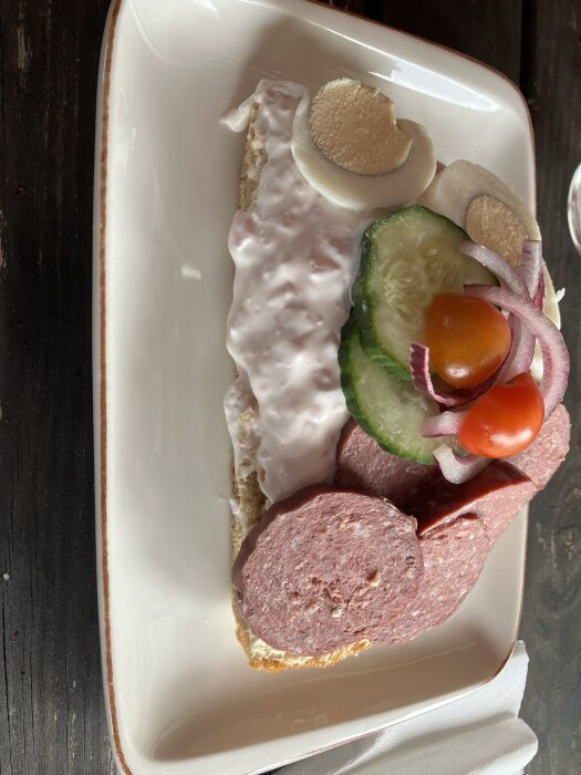 En smörgås med prickig korv, sallad, gurka, ägg och tomater på en vit tallrik på ett mörkt träbord.